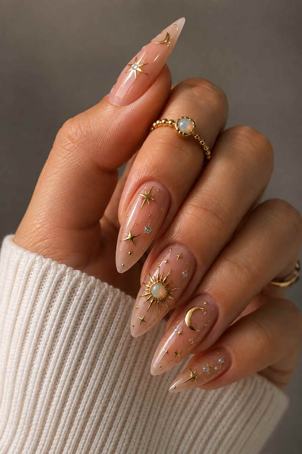 Almond nails with milky pink base, gold star and moon charms, and an opal-centered sun charm