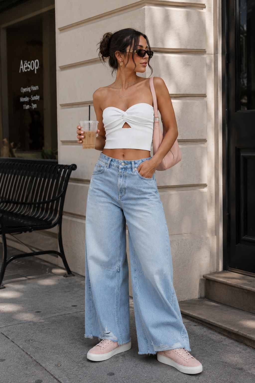Woman wearing a white twist-front crop top, distressed wide-leg jeans, blush pink shoulder bag, and white sneakers.