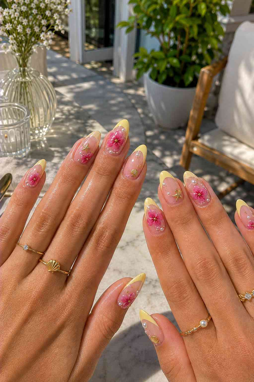 Soft yellow French tip nails with pink flower art, pearls, and small gold starfish and shell charms
